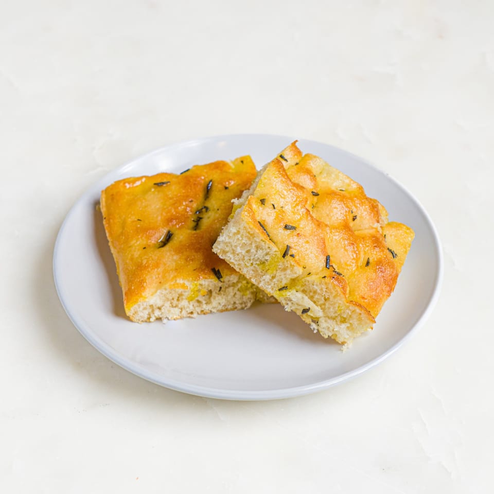 Two golden-brown focaccia bread slices, topped with rosemary and olive oil, on a white plate.